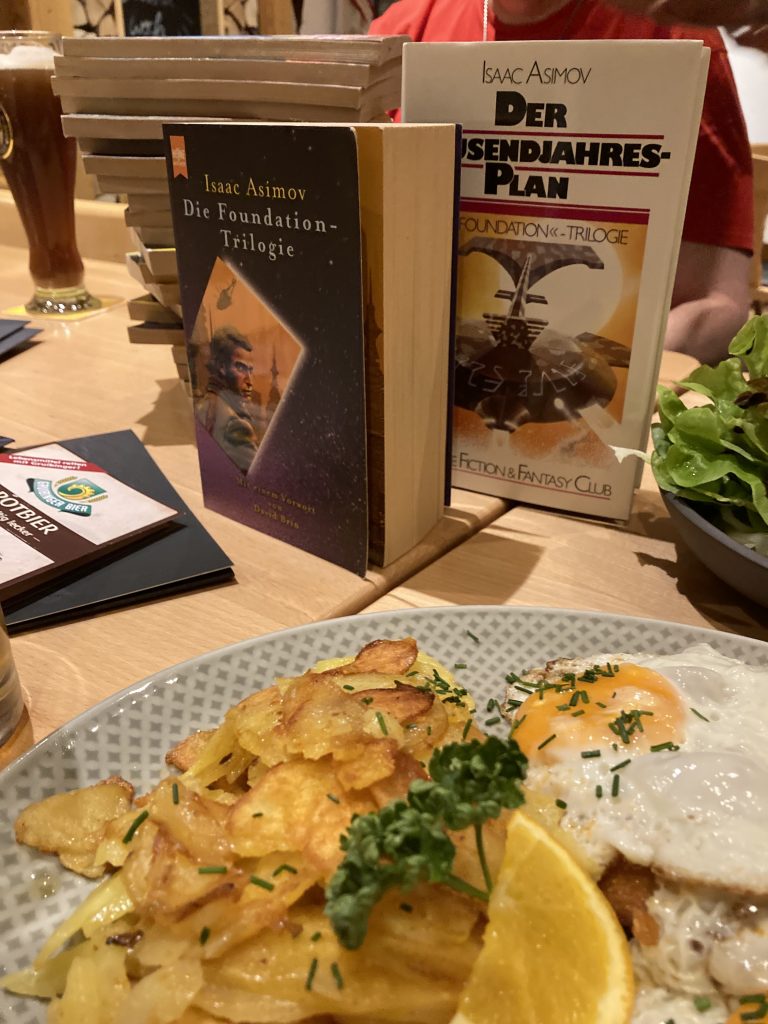 Auf einem Tisch steht ein Teller mit Bauernschnitzel. Dahinter stehen zwei Science Fiction Bücher.